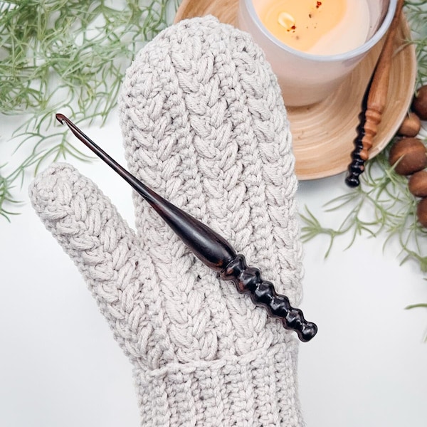 May include: A close-up of a crocheted mitten in a light beige color with a cable knit pattern. A wooden crochet hook is resting on top of the mitten.