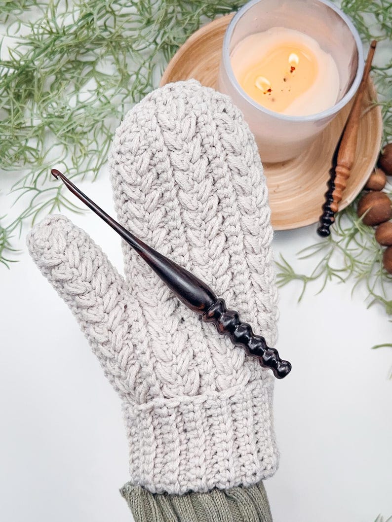 May include: A close-up of a crocheted mitten in a light beige colour with a cable knit pattern. A wooden crochet hook is resting on top of the mitten.
