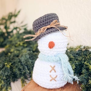 White crochet snowman with gray hat, light blue scarf and twine buttons on a wooden riser with greenery