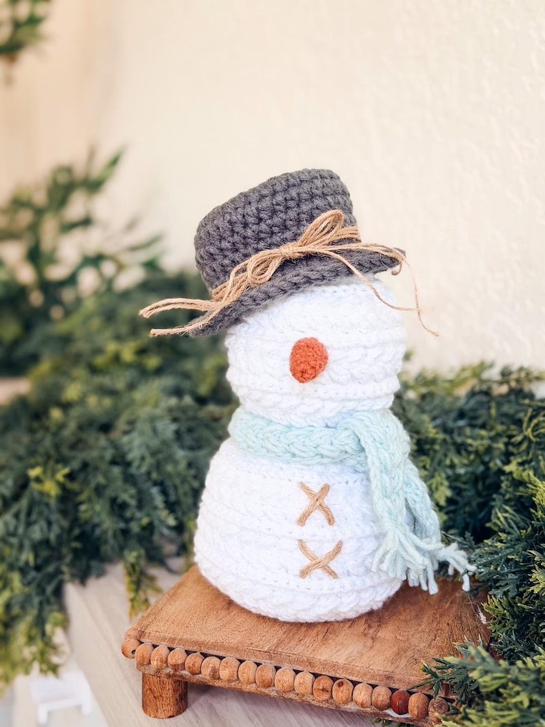 White crochet snowman with gray hat, light blue scarf and twine buttons on a wooden riser with greenery