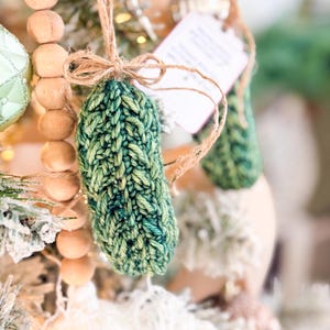 May include: A hand-knitted green pickle ornament made from variegated yarn, featuring a textured stitch pattern. The ornament is suspended by a thin brown twine tied in a bow at the top, with a small white rectangular tag attached to the twine.
