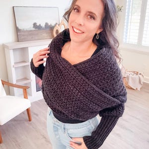 A woman stands indoors wearing a black crochet shawl and jeans, smiling at the camera. A chair, a small shelf with unique jewellery, and a framed picture are in the background.