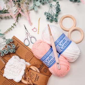 May include: A craft supplies flat lay featuring white and pink Village Yarn Craft Cotton, crochet hooks, scissors, wooden rings, and a handmade white crochet dishcloth. Decorative floral elements surround the items.
