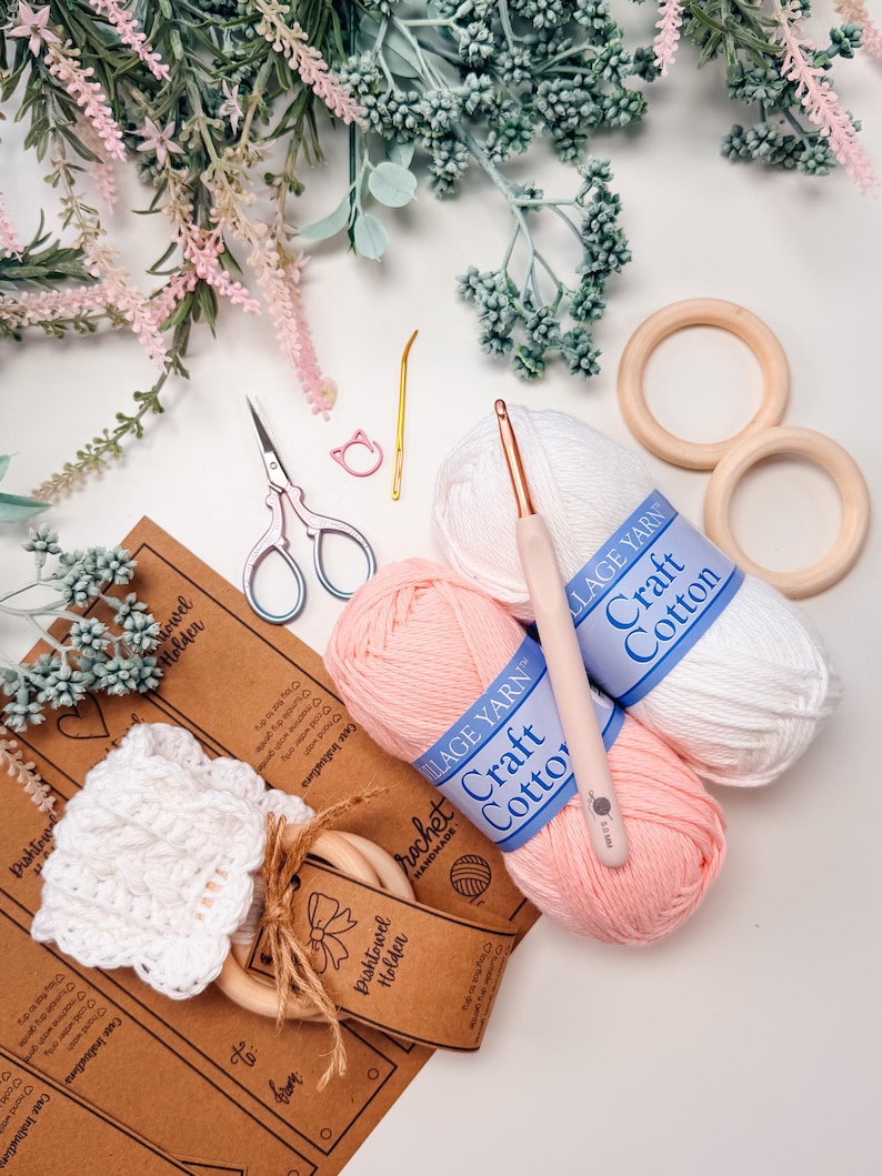 May include: A craft supplies flat lay featuring white and pink Village Yarn Craft Cotton, crochet hooks, scissors, wooden rings, and a handmade white crochet dishcloth. Decorative floral elements surround the items.