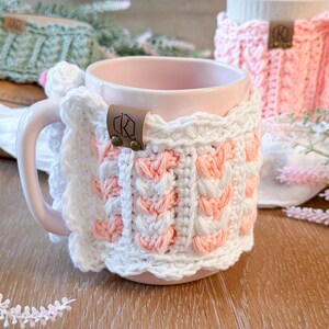 May include: A pink ceramic mug with a white and peach crochet cozy. The cozy features a cable knit design with a scalloped edge and a leather tag. A second mug with a pink cozy is visible in the background.