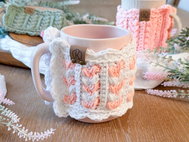 May include: A pink ceramic mug with a white and peach crochet cozy. The cozy features a cable knit design with a scalloped edge and a leather tag. A second mug with a pink cozy is visible in the background.
