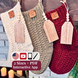 Op de afbeelding: Drie gehaakte kerstsokken in crème, olijfgroen en donkerrood, elk met een gestructureerd kabelbreipatroon, een bruin leren label met een logo en een houten cadeaulabel hangend aan een jute touwtje met drie houten kralen.