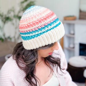 A woman wearing a pastel-colored crocheted beanie and a light pink sweater, paired with unique jewellery, looks down in a bright, cozy room.