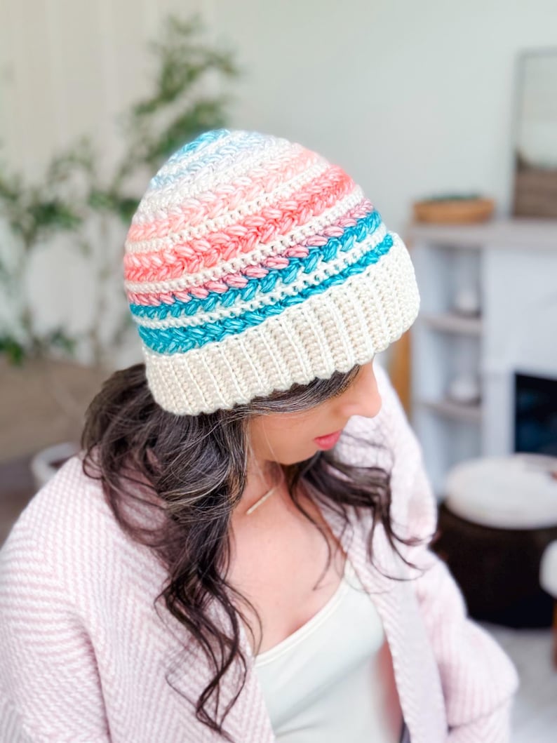 A woman wearing a pastel-colored crocheted beanie and a light pink sweater, paired with unique jewellery, looks down in a bright, cozy room.
