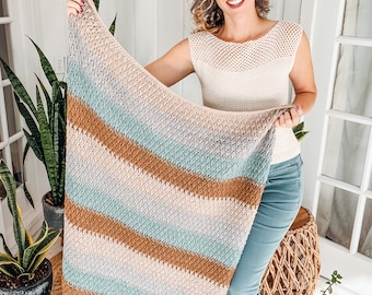 Op de afbeelding: Een rechthoekige gehaakte deken met horizontale strepen in afwisselende kleuren van lichtgrijs, zacht talingblauw en warm bruin. Het textiel heeft over het gehele oppervlak een gestructureerd weefpatroon, wat zorgt voor een consistente uitstraling.