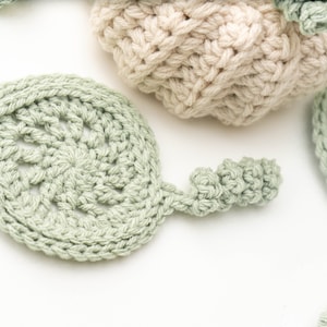 Close-up of a sage green crochet leaf coaster with a lacy center and curly stem near a cream pumpkin