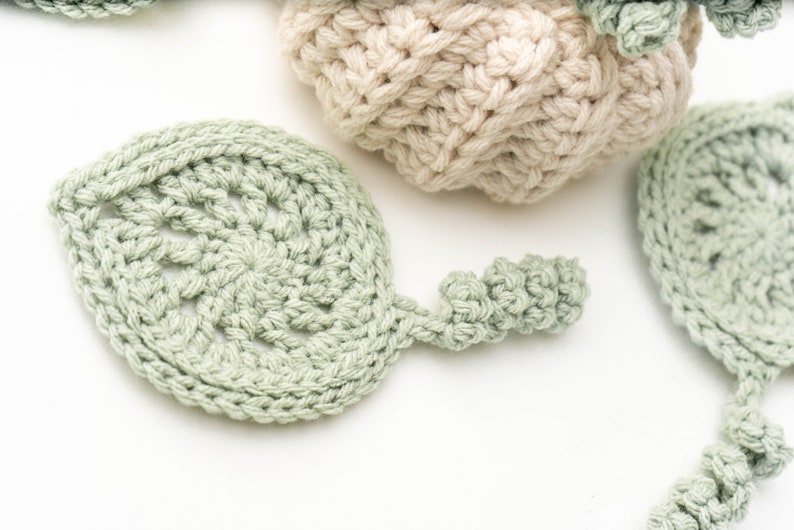 Close-up of a sage green crochet leaf coaster with a lacy center and curly stem near a cream pumpkin