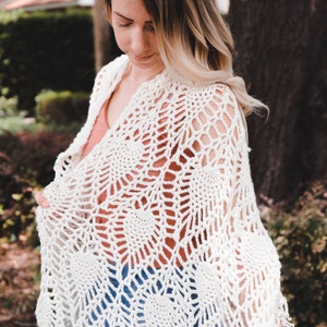 A woman wearing a white shawl decorated with the intricate crochet pineapple stitch, with fringed edges. She stands in an outdoor green space, surrounded by greenery and sunshine.