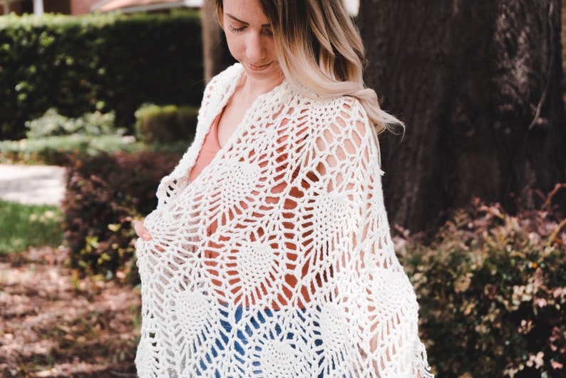 A woman wearing a white shawl decorated with the intricate crochet pineapple stitch, with fringed edges. She stands in an outdoor green space, surrounded by greenery and sunshine.