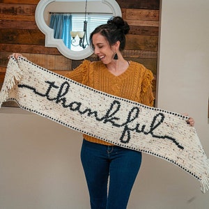 May include: A white crocheted scarf with black yarn spelling out the word "thankful". The scarf has fringe on both ends and is being held by a person wearing a yellow sweater and blue jeans.