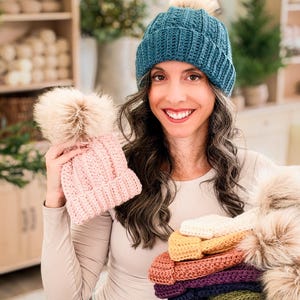 Woman in a teal crochet beanie holds a pink pom pom hat and a stack of colorful beanies