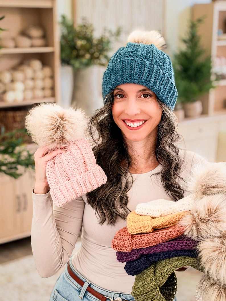 Woman in a teal crochet beanie holds a pink pom pom hat and a stack of colorful beanies