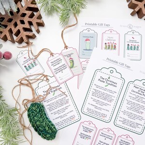 May include: A dark green crocheted pickle ornament with a brown twine loop, accompanied by several printed paper gift tags featuring pickle illustrations and text explaining The Christmas Pickle Tradition, arranged with wooden snowflake decor.