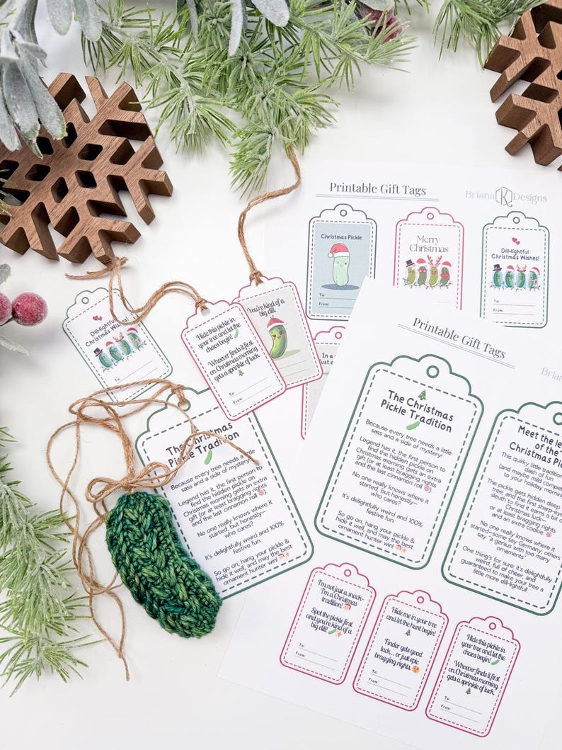 May include: A dark green crocheted pickle ornament with a brown twine loop, accompanied by several printed paper gift tags featuring pickle illustrations and text explaining The Christmas Pickle Tradition, arranged with wooden snowflake decor.