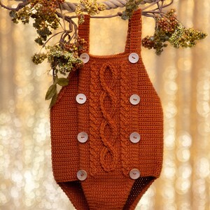 May include: A brown crocheted baby onesie with a cable knit pattern and wooden buttons.