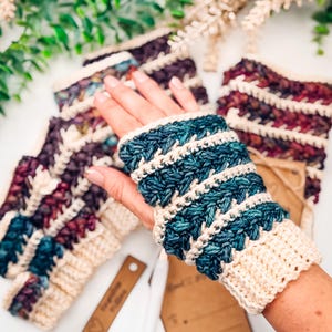 May include: Hand-knitted fingerless gloves in shades of blue, cream, and burgundy. The gloves feature a textured stitch pattern and a cream-coloured cuff. Several pairs are visible, with one worn on a hand.