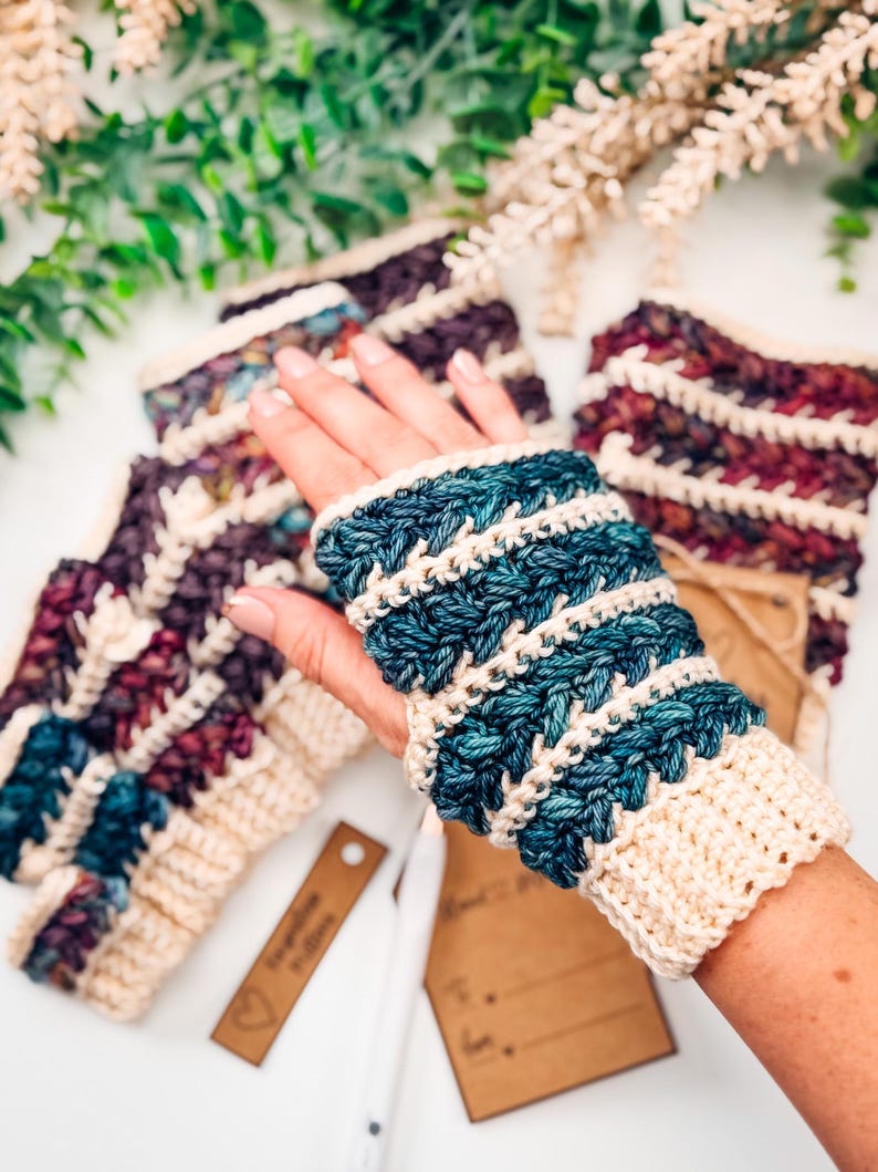 May include: Hand-knitted fingerless gloves in shades of blue, cream, and burgundy. The gloves feature a textured stitch pattern and a cream-coloured cuff. Several pairs are visible, with one worn on a hand.