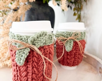 Puede incluir: Dos vasos de café desechables blancos con fundas de ganchillo hechas a mano. Las fundas son de un color terracota intenso con textura acanalada vertical, adornadas con una hoja de ganchillo verde salvia y atadas con un lazo de yute natural.