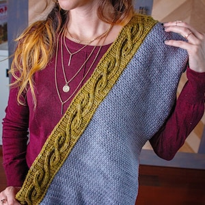May include: A woman wearing a maroon sweater with a gray and yellow crocheted shawl draped over her shoulder. She is wearing a layered gold necklace with a round pendant.