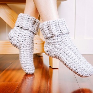 May include: A pair of grey crocheted slipper socks with a folded cuff. The socks are on a wooden surface.