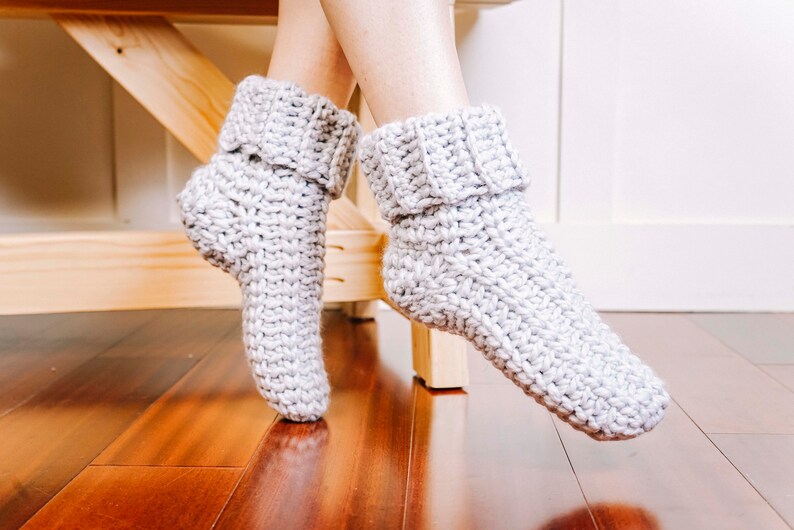 May include: A pair of grey crocheted slipper socks with a folded cuff. The socks are on a wooden surface.