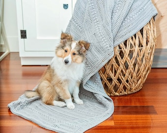 May include: A small, brown and white dog sits on a gray crocheted blanket. The blanket is draped over a wicker basket.