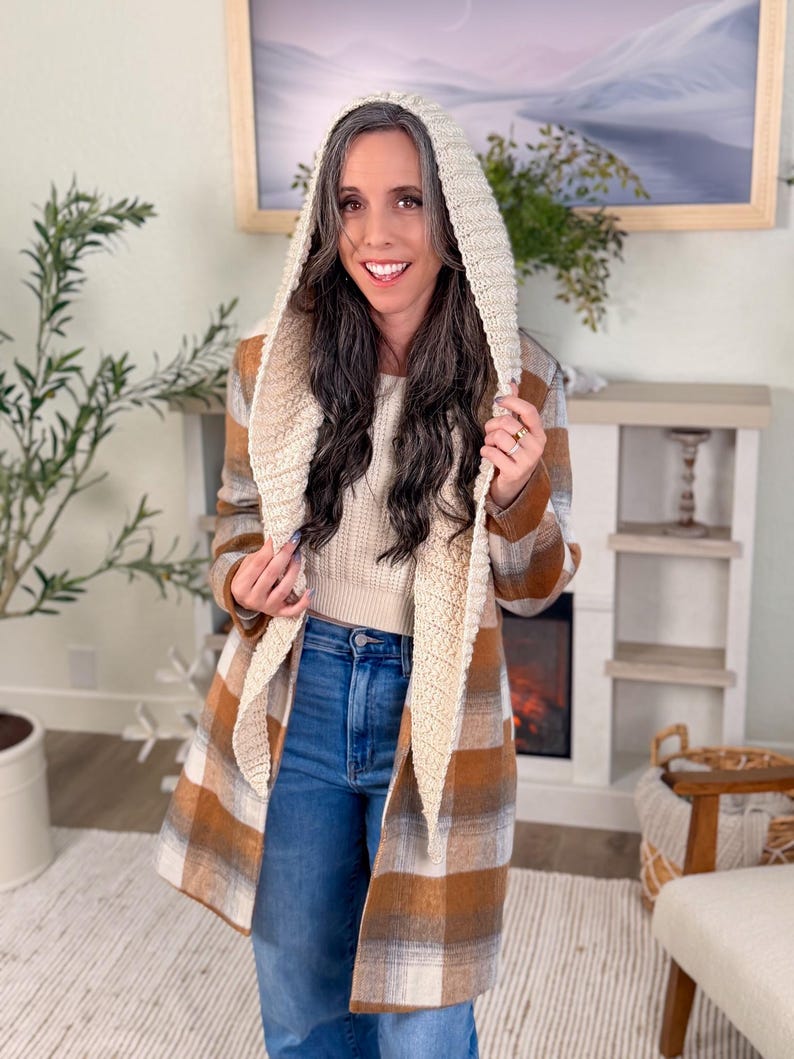 May include: A woman wearing a hooded coat with a plaid pattern in shades of brown and cream. The hood is a cream-coloured knit, and the coat is paired with blue jeans. The outfit suggests a cosy, casual style.