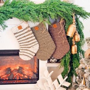 May include: Three handmade crochet Christmas stockings in cream, olive green, and brown hang above a fireplace. The stockings have leather tags and are decorated with a green garland and gold bells. A white snowflake decoration is in the foreground.