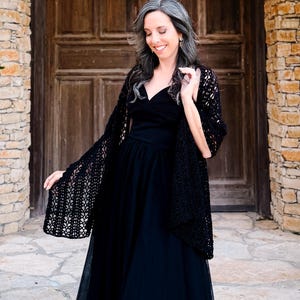 May include: A black sleeveless dress with a sweetheart neckline and a full-length skirt, paired with a black open-front crochet shawl featuring an intricate, repeating leaf-like pattern throughout the fabric and scalloped edges.