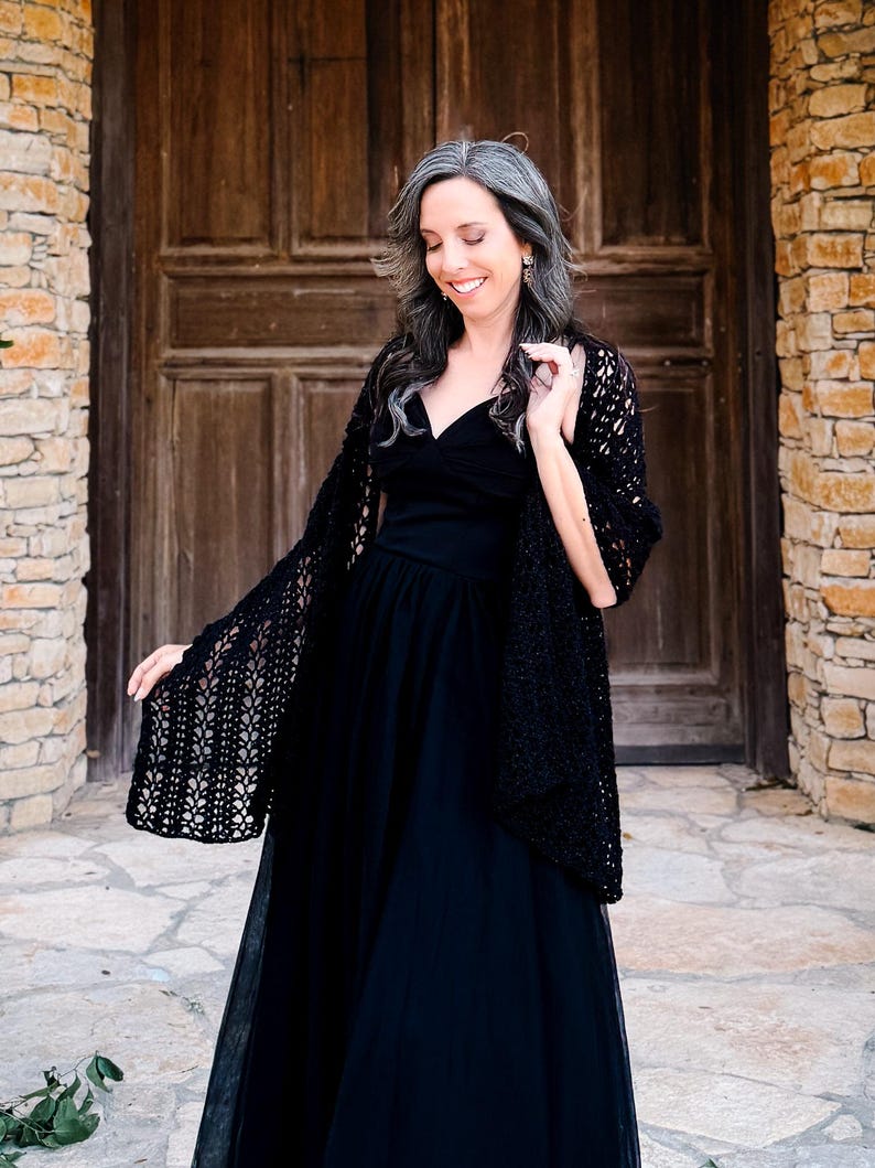 May include: A black sleeveless dress with a sweetheart neckline and a full-length skirt, paired with a black open-front crochet shawl featuring an intricate, repeating leaf-like pattern throughout the fabric and scalloped edges.