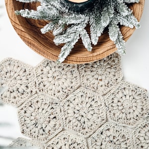 May include: A beige crochet table runner composed of multiple joined hexagonal motifs with a speckled texture. Above the runner sits a round wooden bowl containing a lit candle and a sprig of frosted green pine needles.