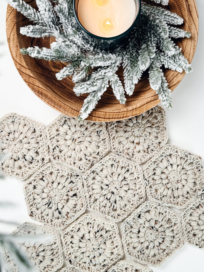 May include: A beige crochet table runner composed of multiple joined hexagonal motifs with a speckled texture. Above the runner sits a round wooden bowl containing a lit candle and a sprig of frosted green pine needles.