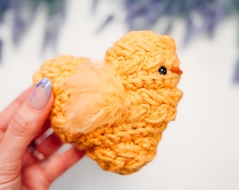 May include: A small, hand-crocheted chick figurine made from bright orange yarn. The bird features a textured, chunky knit body, a single black bead eye, a small orange beak, and a soft, fuzzy wing detail on its side, creating a rounded shape.