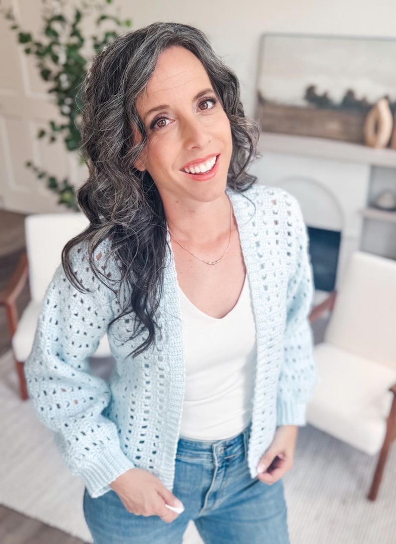 May include: A light blue crochet cardigan with a v-neck white shirt and blue jeans. The cardigan has an open weave design. The person is smiling and wearing a delicate necklace. The background includes white chairs and a fireplace.