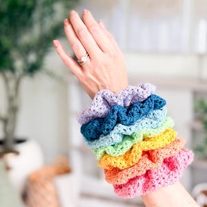 May include: A stack of six crocheted hair scrunchies in a rainbow color gradient, featuring shades of light purple, dark blue, light blue, light green, yellow, orange, and pink, arranged in a vertical row on a wrist.