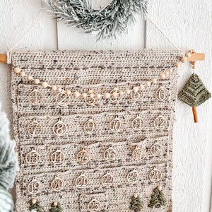 May include: A handmade crochet advent calendar in neutral tones, featuring 25 wooden ornament-shaped tags with numbers. The calendar has small pockets and is adorned with wooden beads and small green crochet Christmas trees. A frosted wreath hangs above.