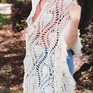 A woman wearing a white shawl decorated with the intricate crochet pineapple stitch, with fringed edges. She stands in an outdoor green space, surrounded by greenery and sunshine.