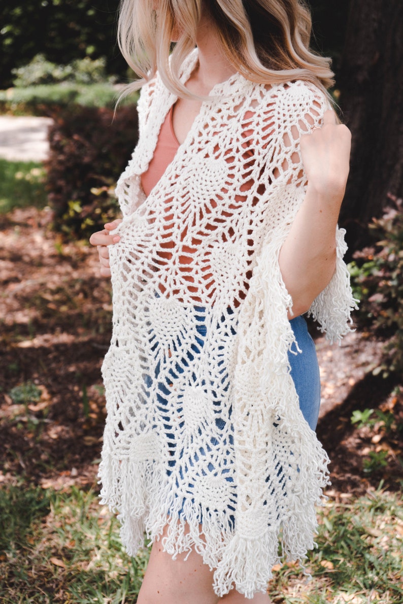 A woman wearing a white shawl decorated with the intricate crochet pineapple stitch, with fringed edges. She stands in an outdoor green space, surrounded by greenery and sunshine.