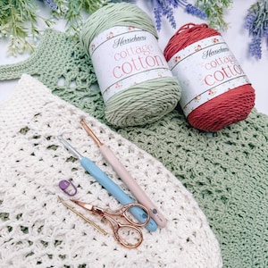 Puede incluir: Una escena artesanal con hilo Herrschners Cottage Cotton en verde y rojo, ganchillos, tijeras y un marcador de puntadas. La escena incluye una pieza de ganchillo blanca y verde, con ramitas de lavanda en el fondo. Las etiquetas del hilo son visibles.