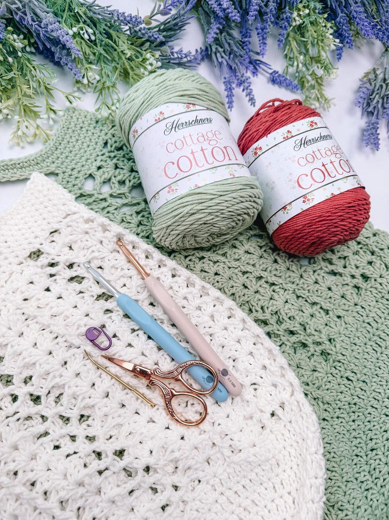 Puede incluir: Una escena artesanal con hilo Herrschners Cottage Cotton en verde y rojo, ganchillos, tijeras y un marcador de puntadas. La escena incluye una pieza de ganchillo blanca y verde, con ramitas de lavanda en el fondo. Las etiquetas del hilo son visibles.