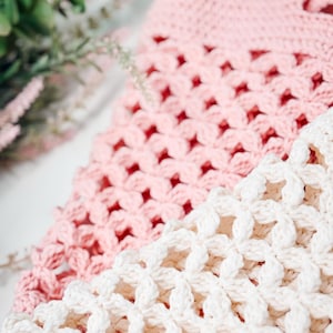 Woman with long brown hair smiles at the camera while holding up a pink crocheted tote bag—a perfect handmade gift—in a well-lit room with plants in the background.
