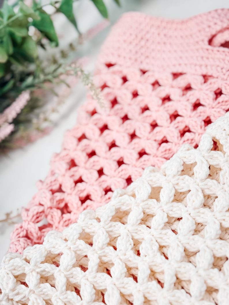 Woman with long brown hair smiles at the camera while holding up a pink crocheted tote bag—a perfect handmade gift—in a well-lit room with plants in the background.