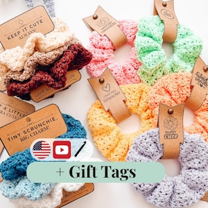 Puede incluir: Una colección de coleteros de ganchillo hechos a mano y coloridos en varios tonos, incluyendo rosa, verde, amarillo y azul. Cada coletero se presenta con una etiqueta de regalo marrón. Las etiquetas presentan textos como "Keep it Cute Always!" y "Tiny Scrunchie, Big Charm!"