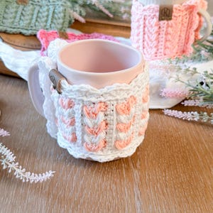 May include: A collection of handmade crochet mug cozies in various colors, including white, pink, and green. The cozies are wrapped around ceramic mugs, showcasing the intricate stitch patterns and textures. Each cozy features a small leather tag.