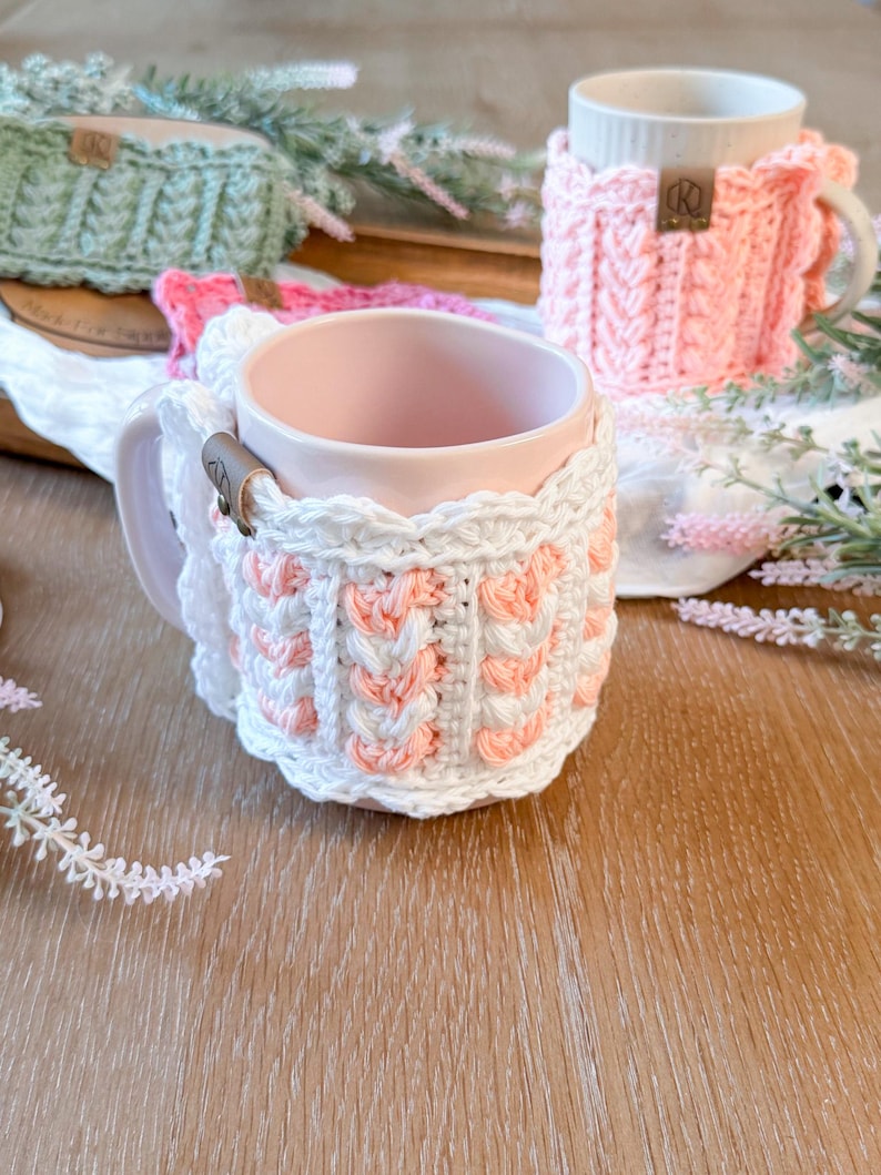 May include: A collection of handmade crochet mug cozies in various colors, including white, pink, and green. The cozies are wrapped around ceramic mugs, showcasing the intricate stitch patterns and textures. Each cozy features a small leather tag.